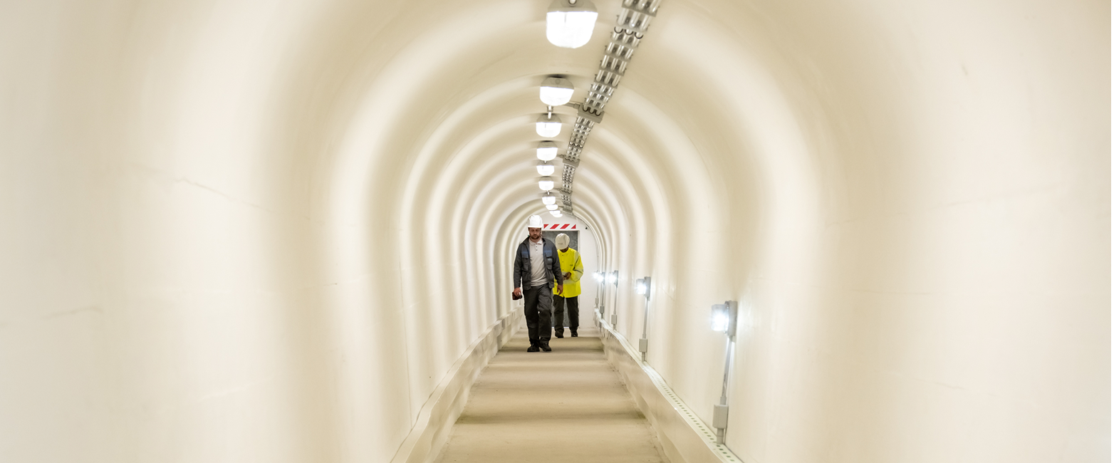 Due tecnici Acea Ato 2 in abiti da lavoro camminano lungo un corridoio dalle pareti bianche nell'infrastruttura del sistema acquedottistico del Peschiera-Capore